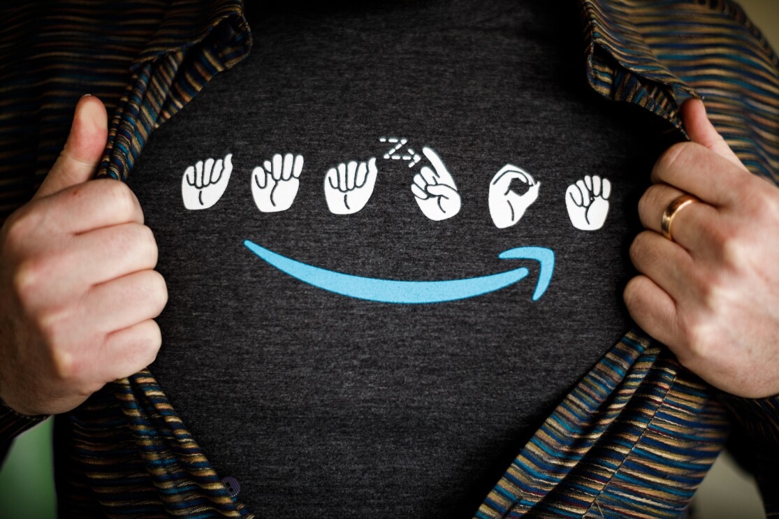 A man wears a t-shirt with the Amazon logo in sign language. Under the signs is a blue Amazon smile. The man is wearing a striped button down shirt over the t-shirt, and is holding the edges of his button down to make the entire logo visible.