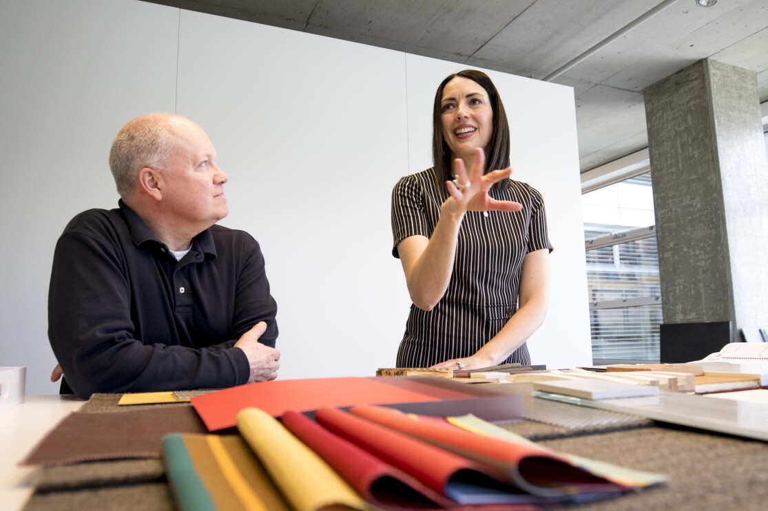 A woman in a grey-striped shirt stands over fabric swatches, talking with a male colleague, her right hand extended in front of her.
