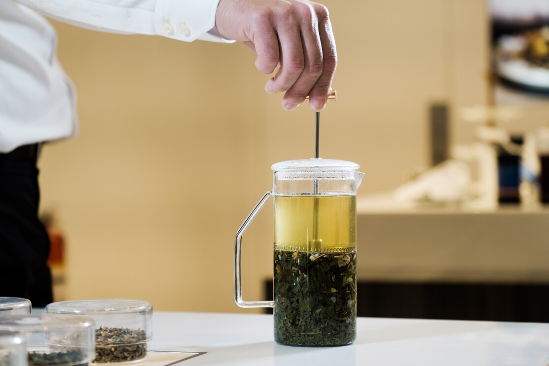 Tea steeps in a French press style pitcher. A person's hand is poised atop the plunger that is pressed down to isolate the tea leaves from the finished beverage.