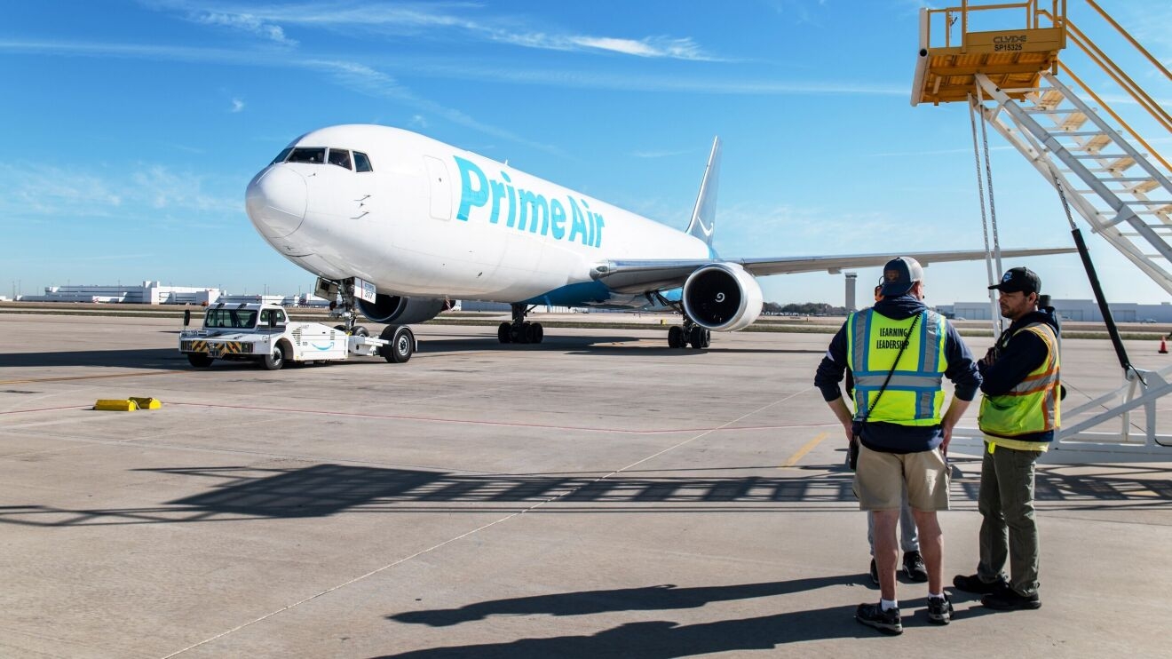 An image of an Amazon Air plane parked outside on an air hub. There is a small car pushing the plane back onto the taxiway. 