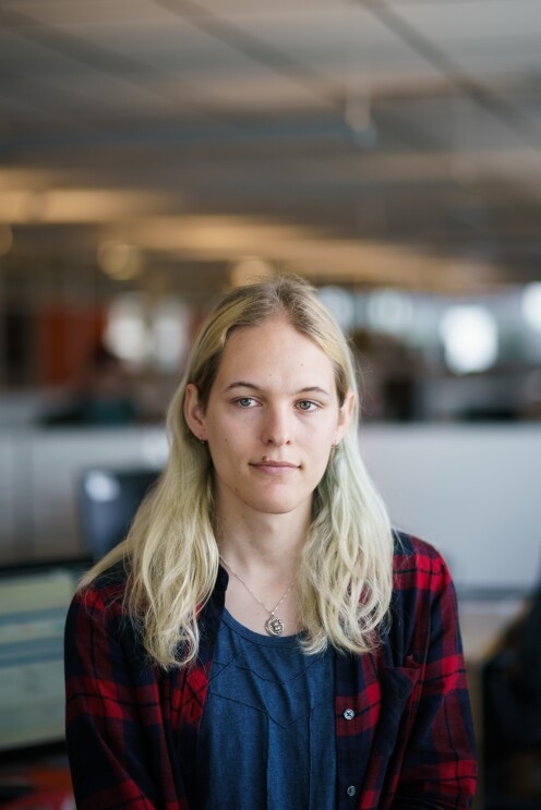 Danielle Skysdottir in an office space with cubicles in the background. She has long blond hair and wears a T-shirt, a flannel shirt, and a necklace with a pendant.