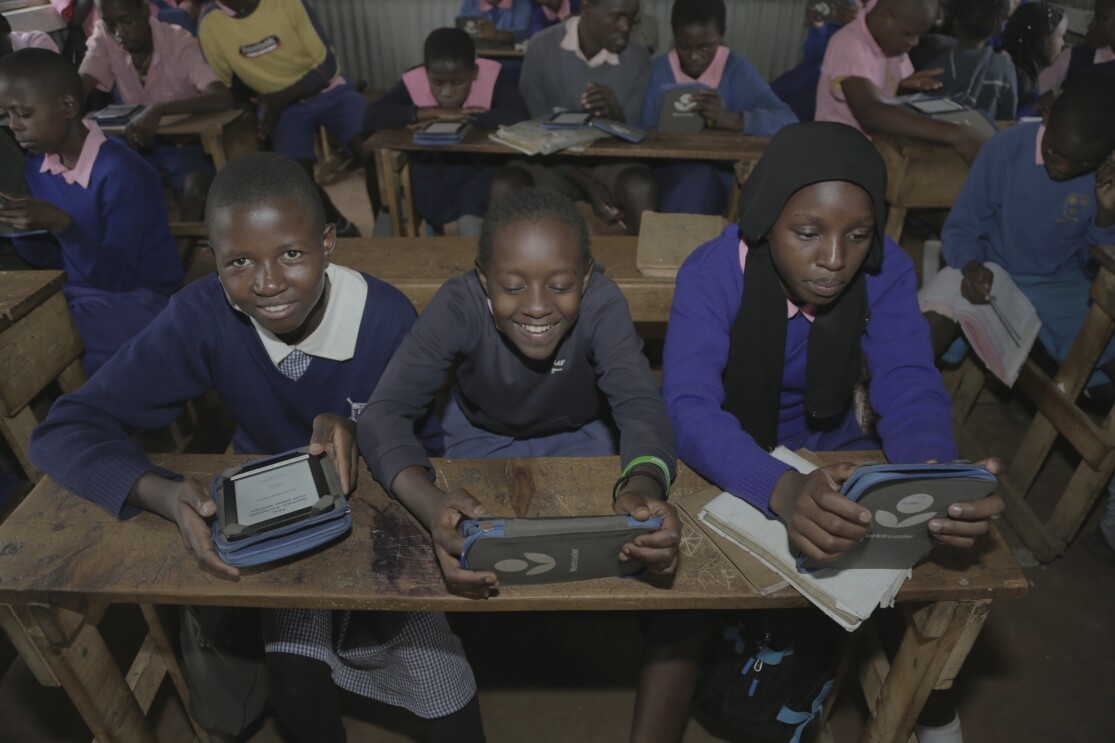 Three girls in blue school uniforms read Kindle Worldreaders at a wooden desk.