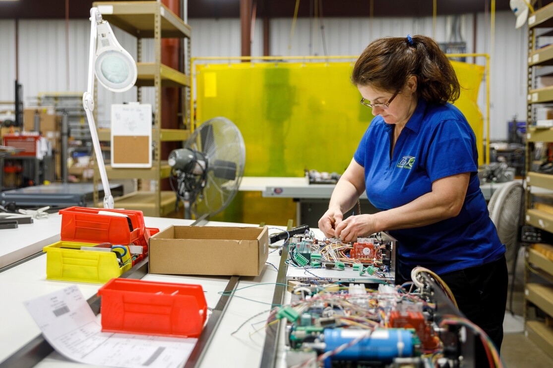 A woman in a blue polo shirt and black pants stands at a work table and assembles electronics.