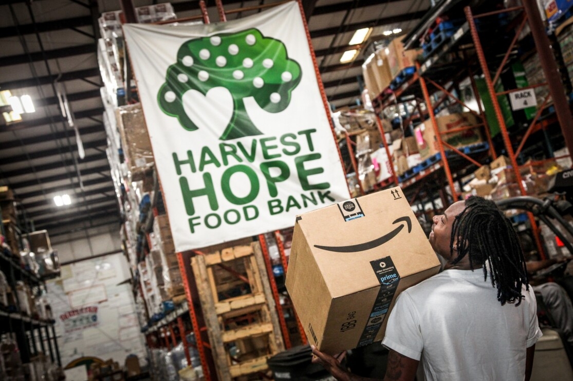 A man walks through a warehouse space under a banner marked "Harvest Hope Food Bank." He carries an Amazon box.