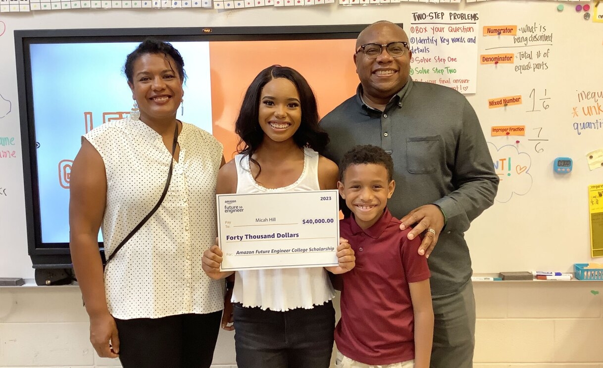 An image of Micah Hill, a 2023 Amazon Future Engineer Scholarship recipient, smiling at the camera with her family and holding out the fund check.
