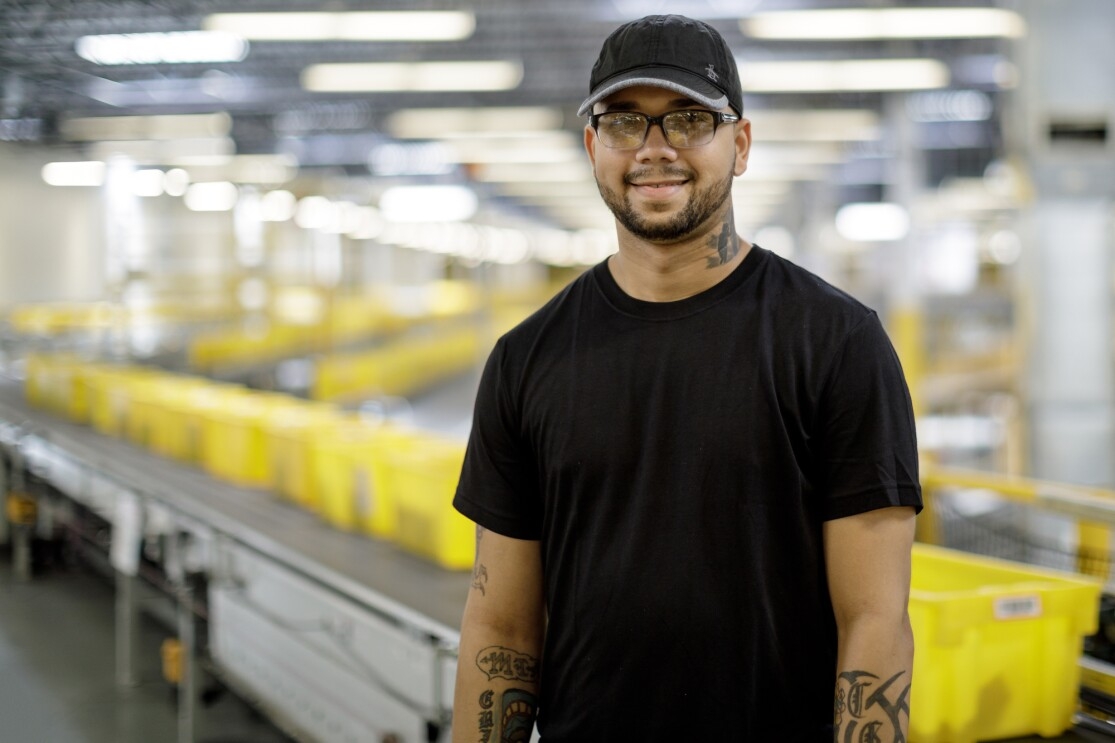 A man stands in a  large space flanked by a conveyor belt carrying yellow tote boxes. He wears glasses, a baseball cap, a black T-shirt, and work gloves.