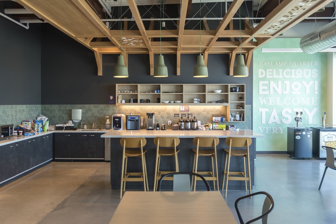 The kitchen area of an Amazon office building in San Diego. Open ceilings and industrial lighting are over an island. In the background are coffee makers, water machines and other kitchen accessories.