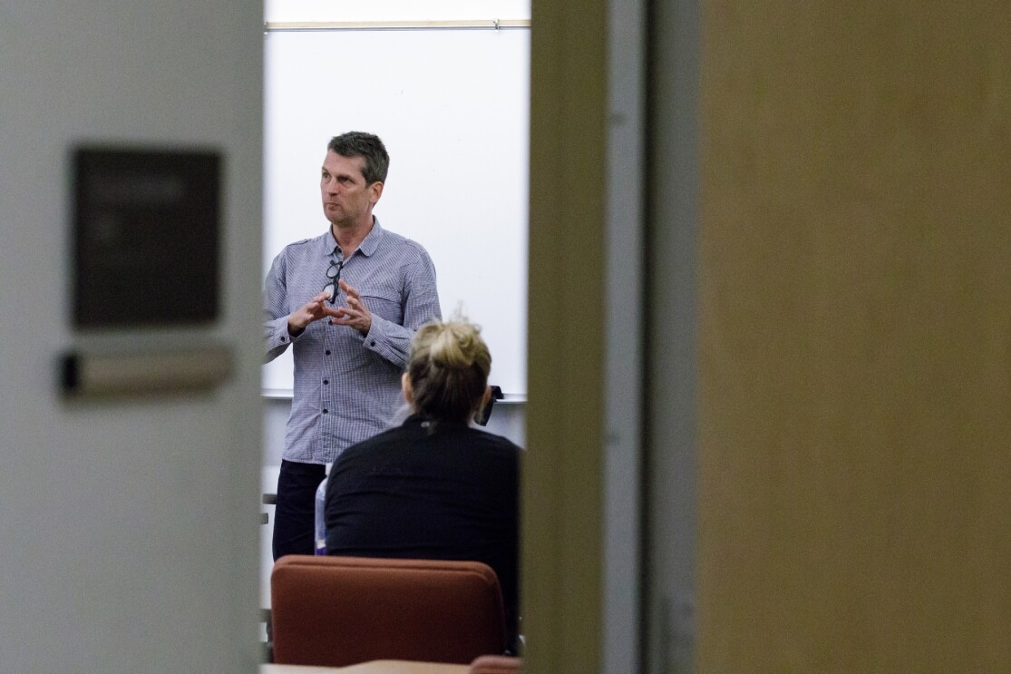 Man in a button-down shirt stands at the front of a classroom. In the foreground, the doorway of the classroom is visible.