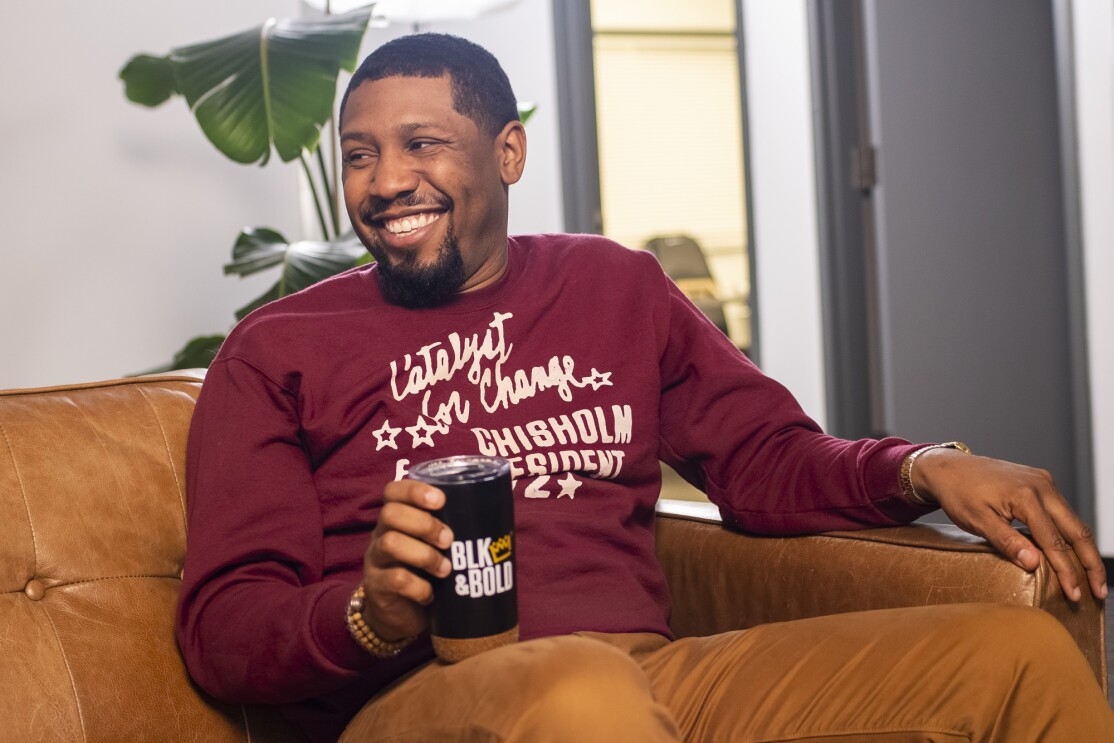 A smiling man sits on a couch and holds a coffee tumbler.