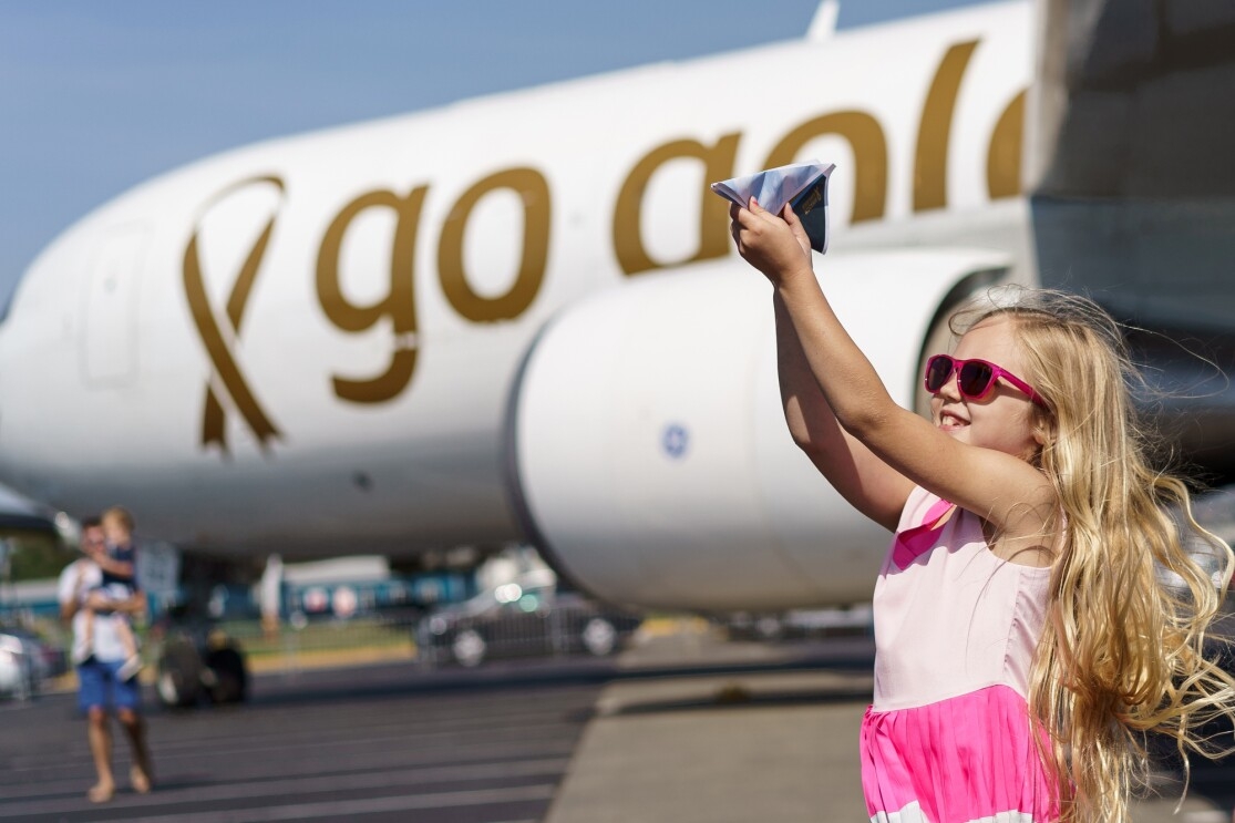 An aircraft with custom messaging applied to it, stating "go gold ...because kids can't fight cancer alone." In front of the airplane, is a young girl with long blonde hair, pink sunglasses and a pink dress flying a paper airplane.