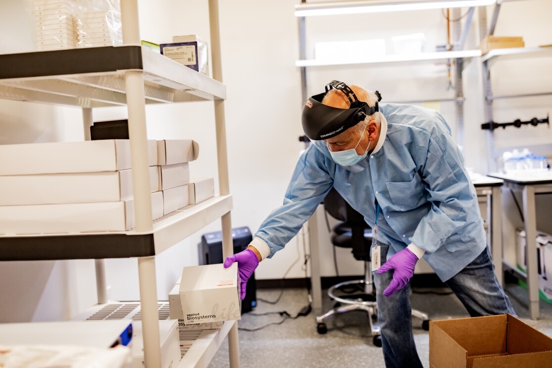 A man wearing protective gear, including a mask, gloves, and face shield, places a box onto a shelf in a laboratory setting. 