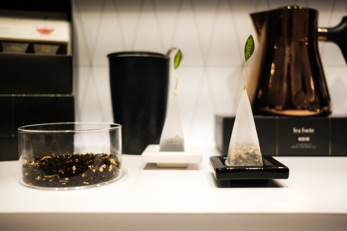 Loose tea leaves in a clear glass container next to two pyramid-shaped teabags.