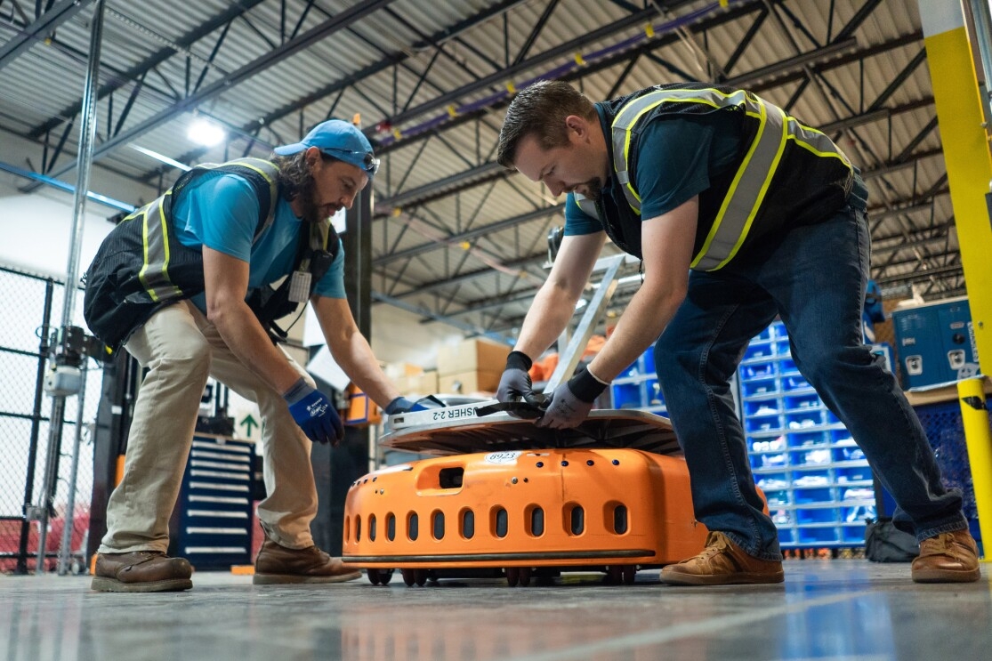 Two men in a warehouse space reach down to secure a silver-colored object to an orange, wheeled piece of machinery.