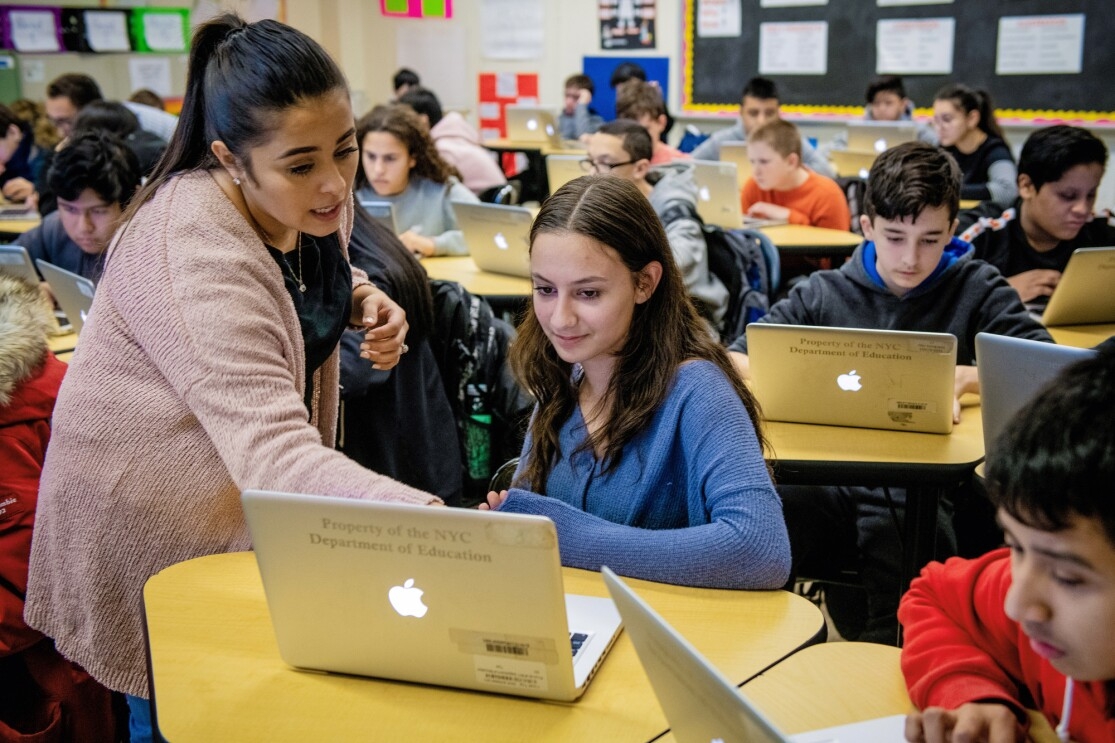 A woman leans over a student's laptop computer, as she explains something to the student. Around her are other students at their desks, working on their own laptops.