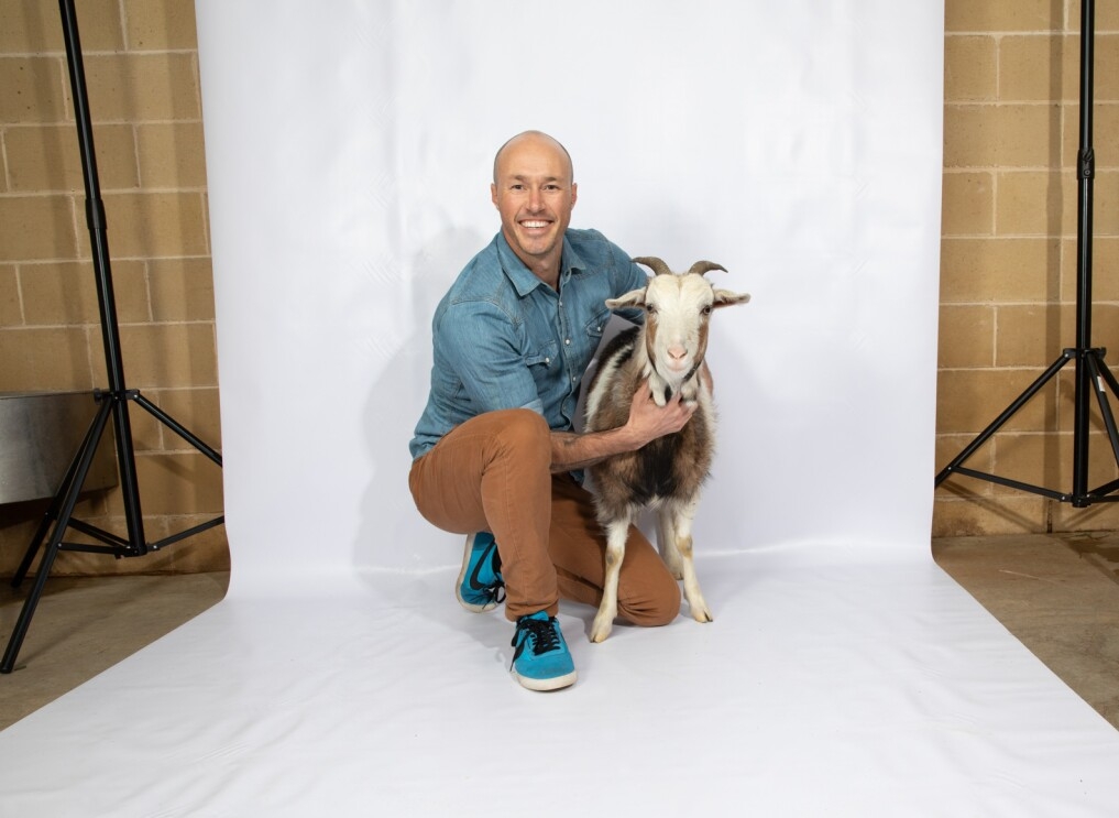 A man in a denim shirt and khaki pants with tennis shoes crouches on a white background. To his left is a small goat, with which he is posing. Both the man and goat are looking into the camera.