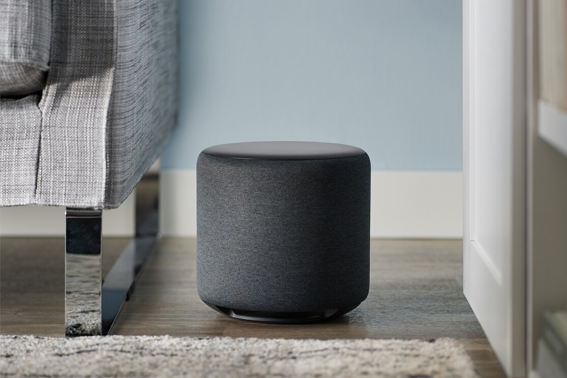 An Echo Sub sits on a wood floor in a living room. To the left is a sofa. To the right, cabinets.