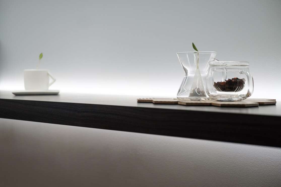 Three pieces of teaware sitting on a shelf. On the left is a white teacup with a cover adorned with a green leaf. On the  right are two glass teapots.