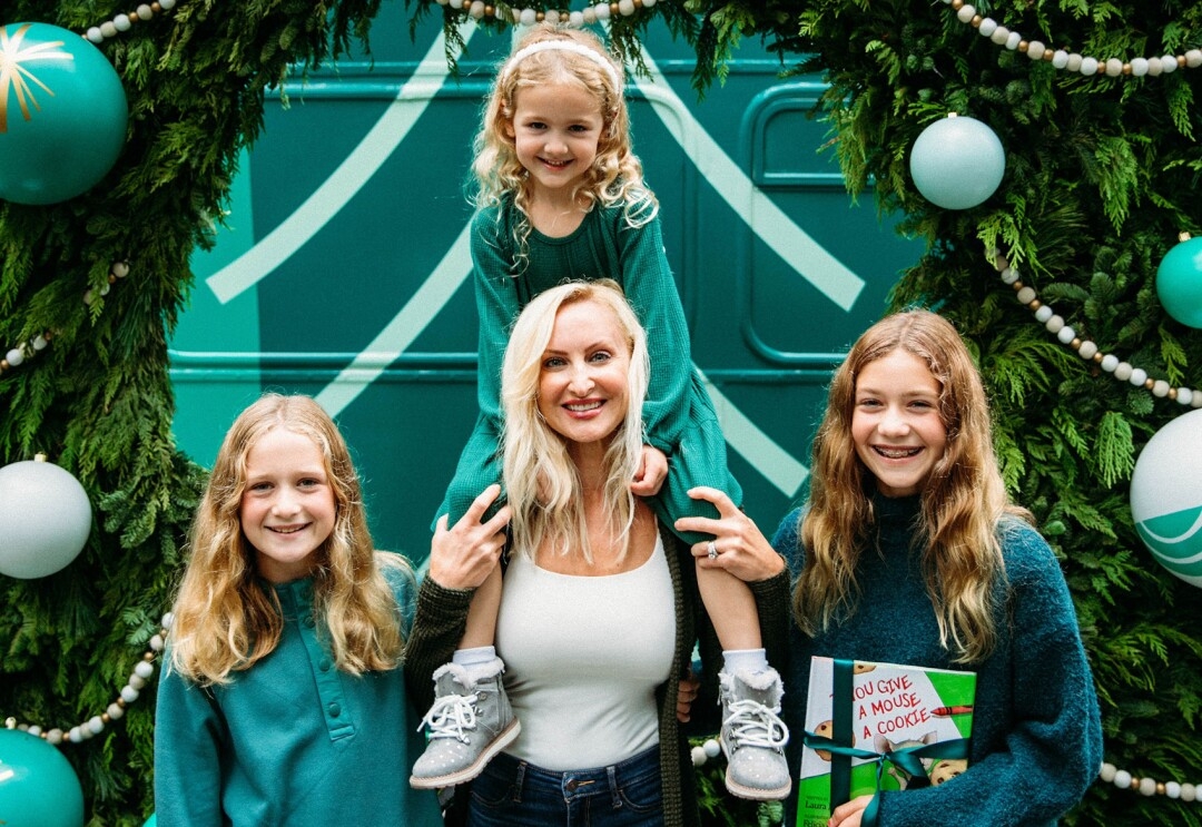 An image of a family smiling for a photo in front of a wreath. The youngest daughter is sitting atop her mother's shoulders. 