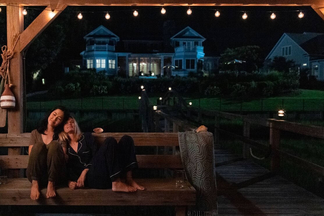 An image of two friends sitting close to each other on a bench on a boat dock at night. One of the women has her head on the other's shoulder and they're smiling and looking off into the distance. 