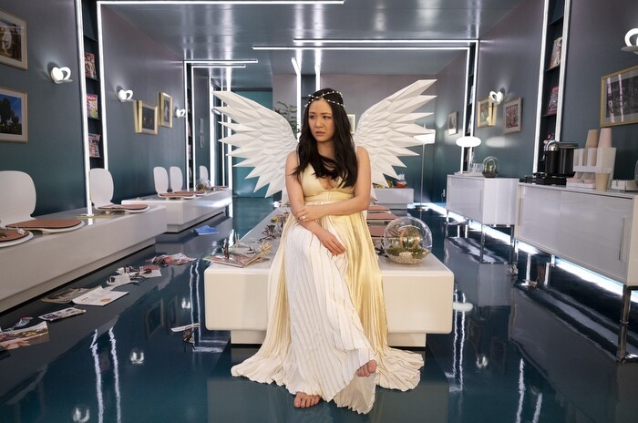 An image of Constance Wu sitting on a table in a clinical waiting room. She is wearing a long, white dress, angel wings, and a small, gold crown. 