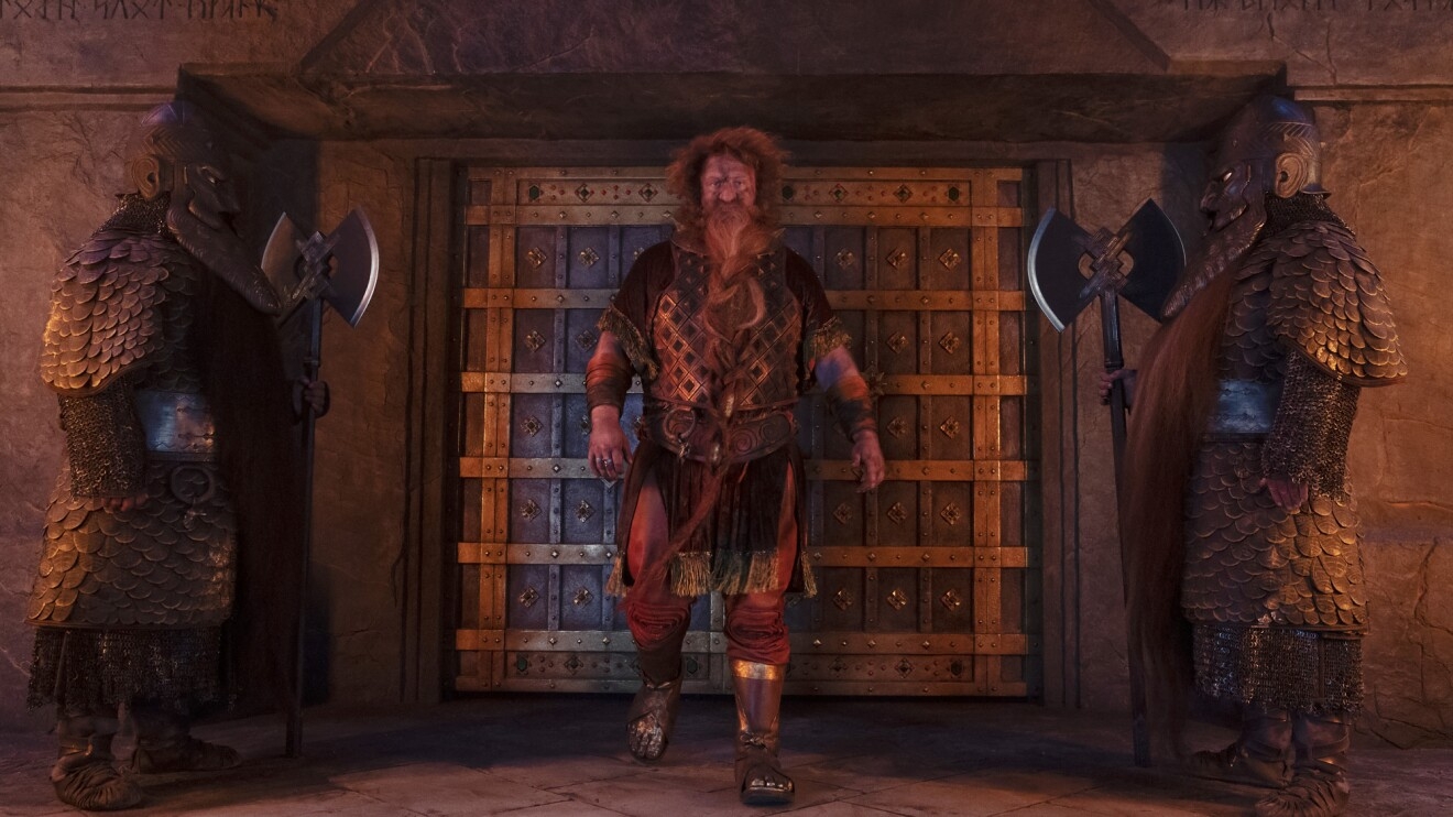 A dwarf-like character takes a step toward the camera as two guards in armor face him from either side of a door.