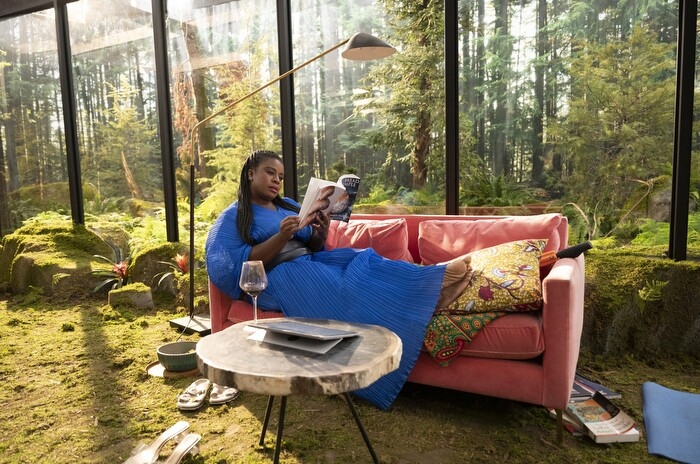 An image of Uzo Aduba sitting on a coach in a home that has large windows showing a green landscape outside and green grass coming through the home as carpet. She is wearing a bright blue dress that contrasts with the green.