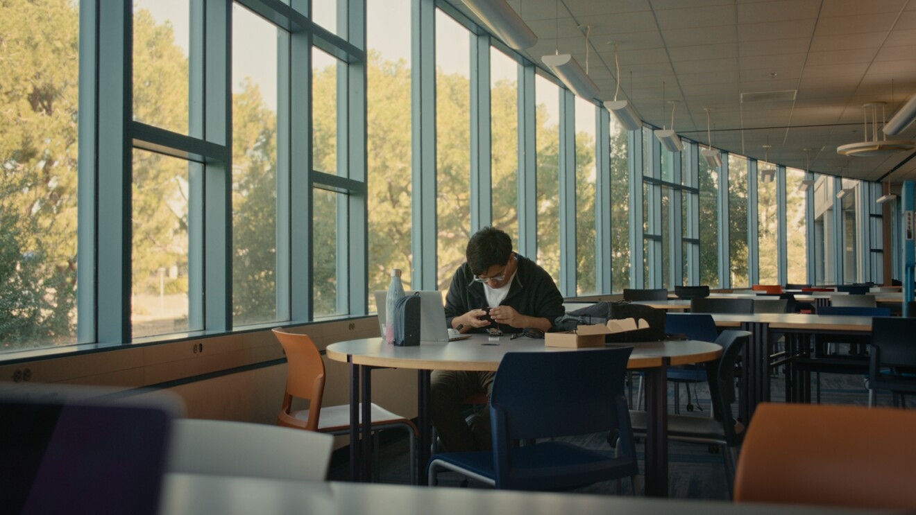 A photo of José Tapia working on a machine at a table inside a community space at California State University, Fullerton.