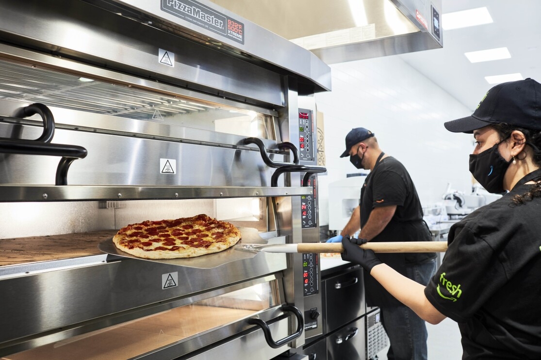 An image of Amazon Fresh employees pulling a fresh pizza out of an oven.