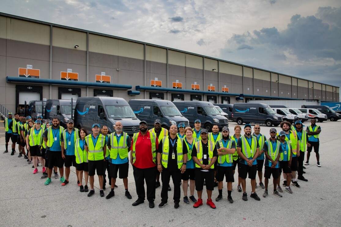 A group of employees stand in front of a fleet of Amazon Prime-branded cargo vans.