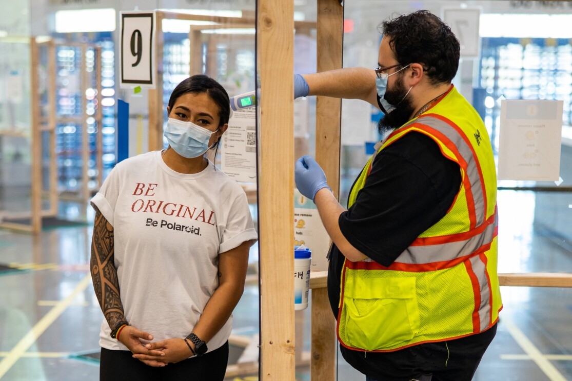 A man in a mask and safety vest reaches through an opening in a see-through barrier and uses a thermometer to take the temperature of a woman wearing a mask.