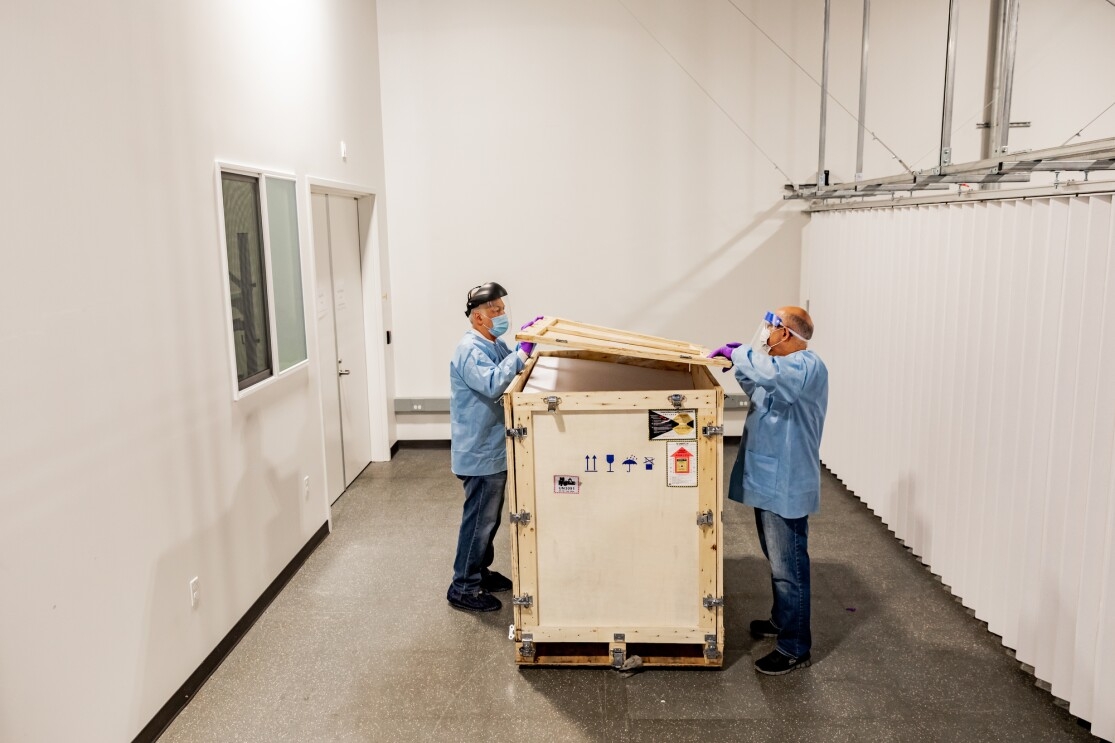 Two men wearing protective clothing, face shield, face mask, and gloves open a large wooden crate. 