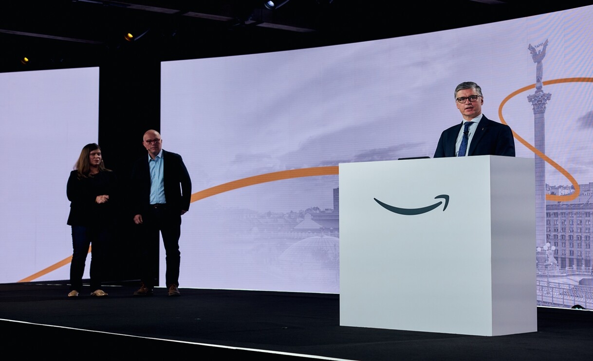 Ukraine’s Ambassador to the UK Vadym Prystaiko stands behind a white podium with the Amazon smile logo on it as he addresses Amazon employees. Liam Maxwell, director of the Government Transformation team in the AWS public sector, and AWS Director of Global Social Impact Maggie Carter stand on the stage listening to Prystaiko speak.