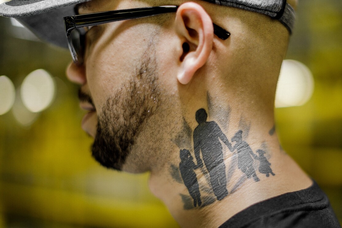 A man wearing glasses is pictured in profile from the shoulders up. He wears glasses and has a beard. A tattoo covers much of the left side of his neck. It shows an adult figure holding hands with three children. All the figures are in silhouette.