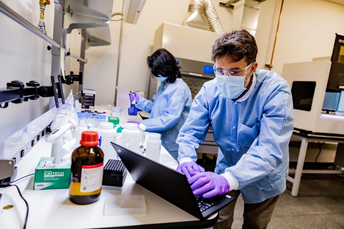 A man in protective gear including safety glasses, mask, gloves, and protective clothing works on a laptop in a laboratory setting. 