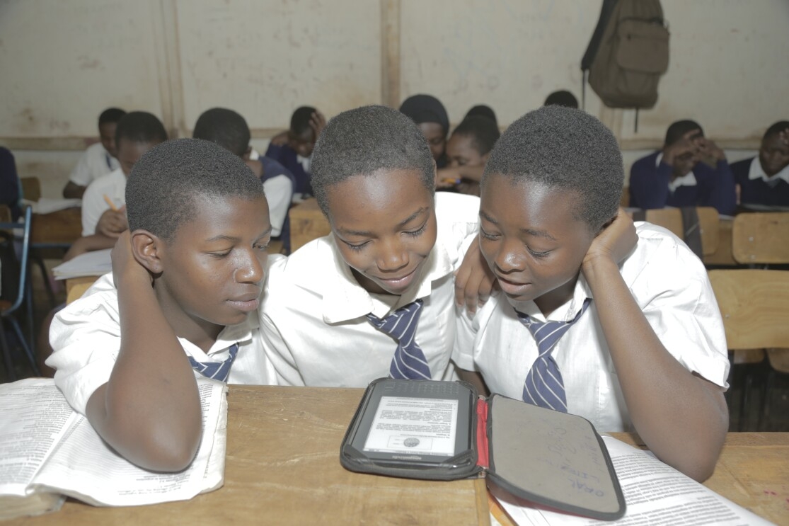 Three boys in school uniforms of white shirts and blue and white striped ties read a Kindle Worldreader.