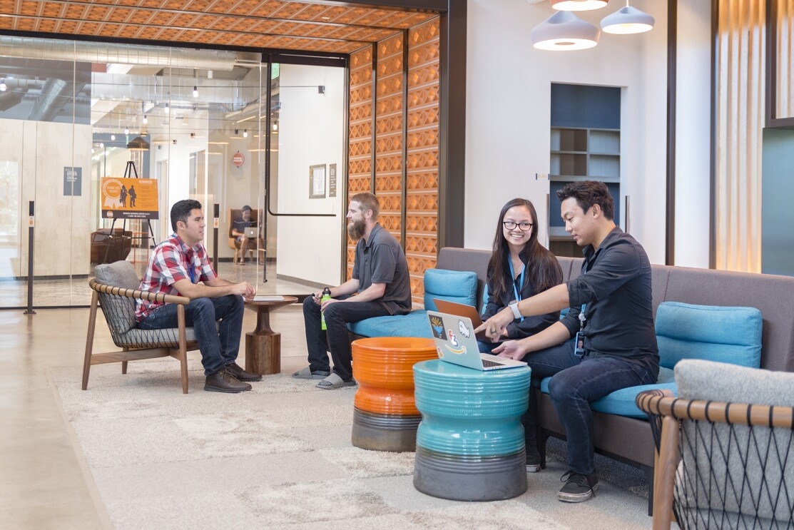 Two men sit at a table in a casual office environment. Near them, a man points at his computer while a female coworker looks at his work.