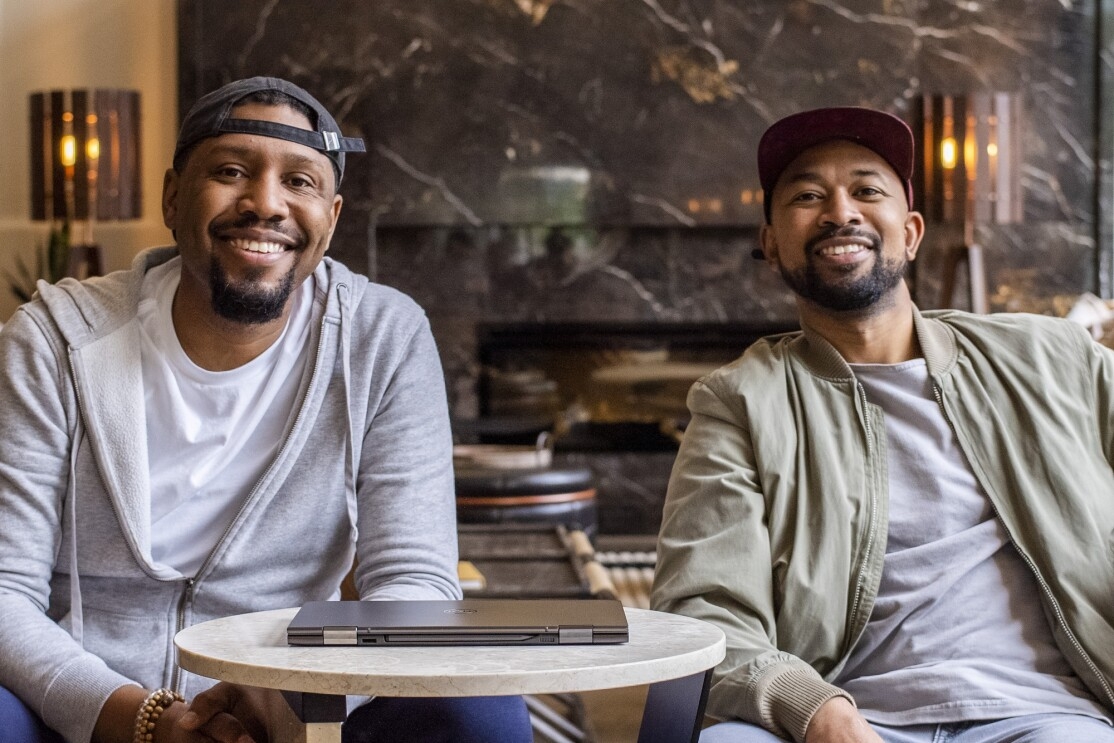 Two smiling me sit next to each other with a closed laptop computer.