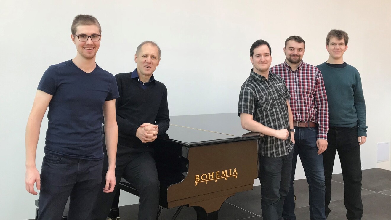 A group of four students and their advisor stand near a Bohemia piano.