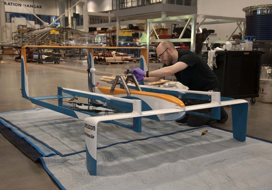 Amazon Prime Air drone in foreground. Behind the drone, a man is kneeling as he appears to be working on the drone. Near the drone, tools are seen.