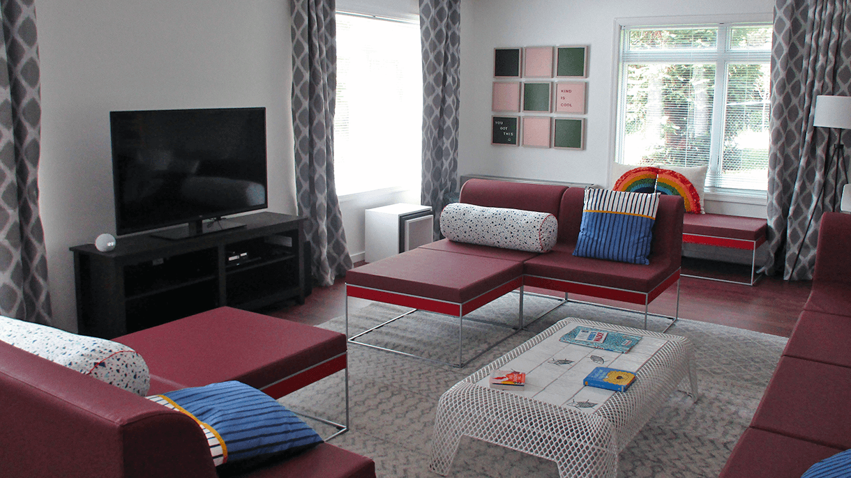 The living room at the Ronald McDonald House in Washington D.C. features a room filled with couches and fun throw pillows. There is a white coffee table with games on top and an Amazon Echo Dot sits next to a TV.