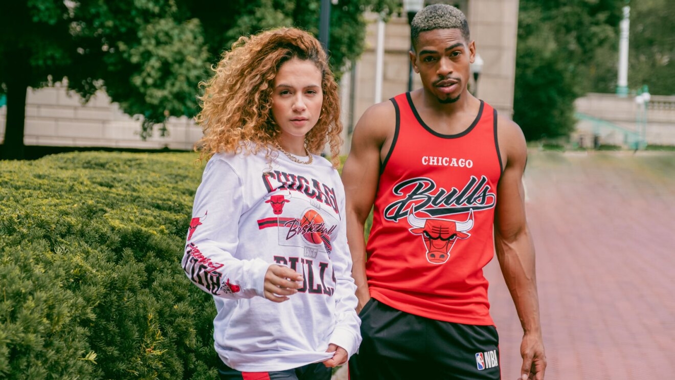 An image of a man and a woman standing on the sidewalk in front of a row of bushes. The woman is wearing a white, long-sleeved shirt that says "Chicago Bulls" in red and black lettering. The man is wearing a red tank top that says "Bulls" in large cursive letters with the Chicago Bulls logo below it.