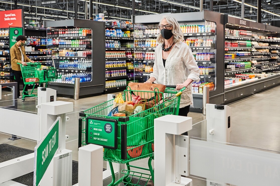 An image of a woman pushing a shopping cart out of the Just Walk Out gates in an Amazon Fresh store. Her cart is full of groceries purchased using the Just Walk Out technology.