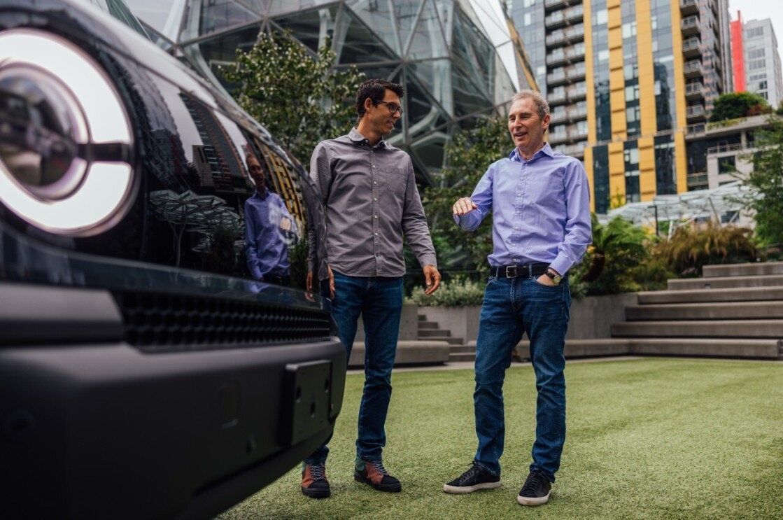 Andy Jassy and RJ Scaringe stand and chat in front of a Rivian Amazon delivery vehicle that is parked outside the Spheres at Amazon headquarters.  