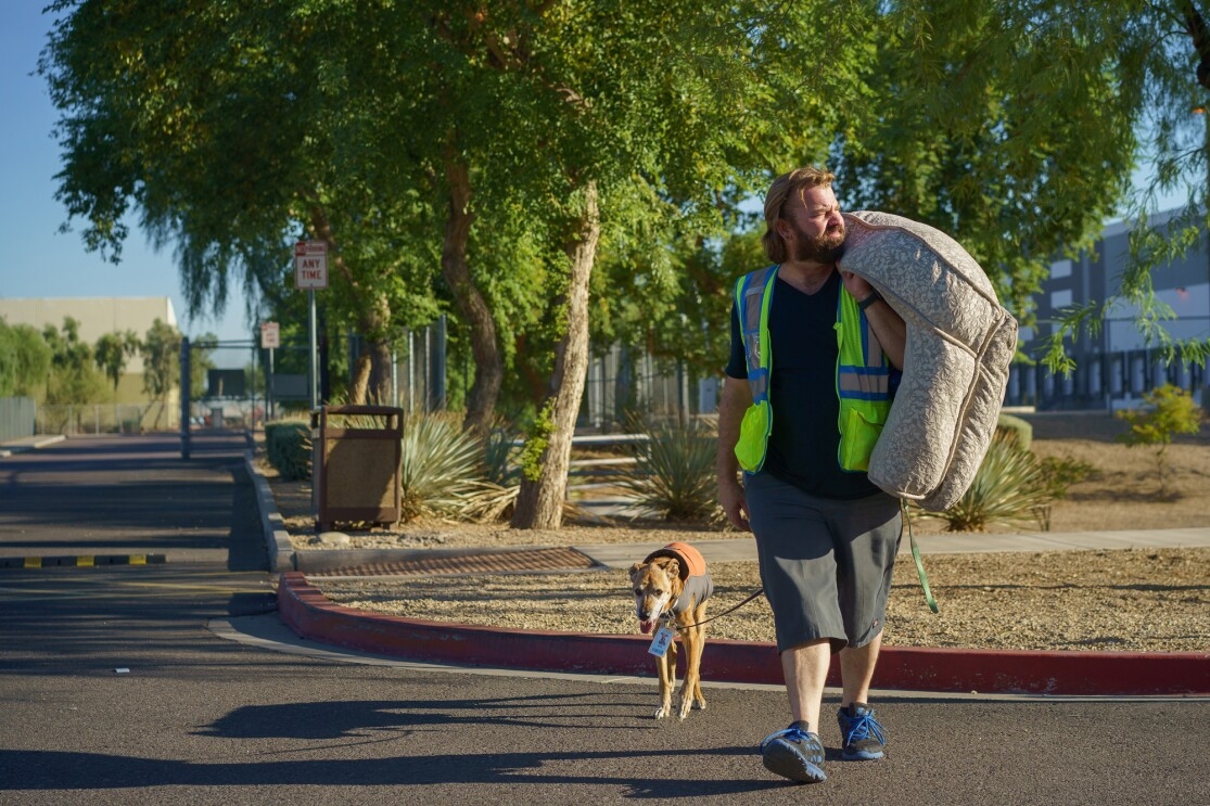 A dog carries a dog bed and leads a dog through a parking lot.