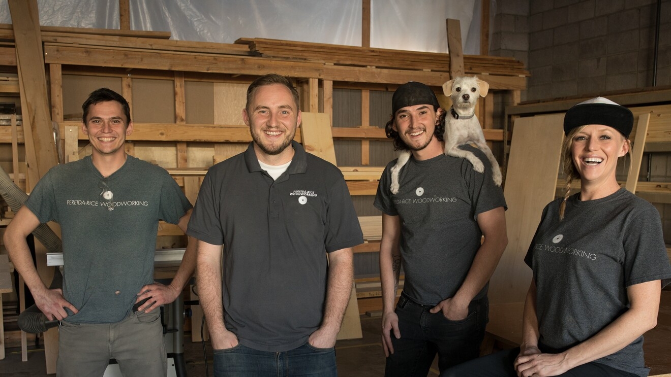 Three males and a female stand in front of a wall of wood. One of the men has a dog perched on his shoulders. These four individuals are siblings who run a small business named Pereida-Rice Woodworking, which sells on Amazon.