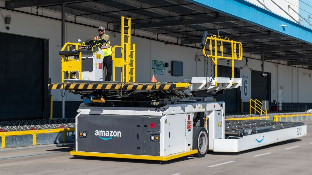 A large vehicle outside of the Amazon Air Hub. There is someone steering the vehicle from a cockpit station. The vehicle is used to carry big, metal containers carrying Amazon packages.