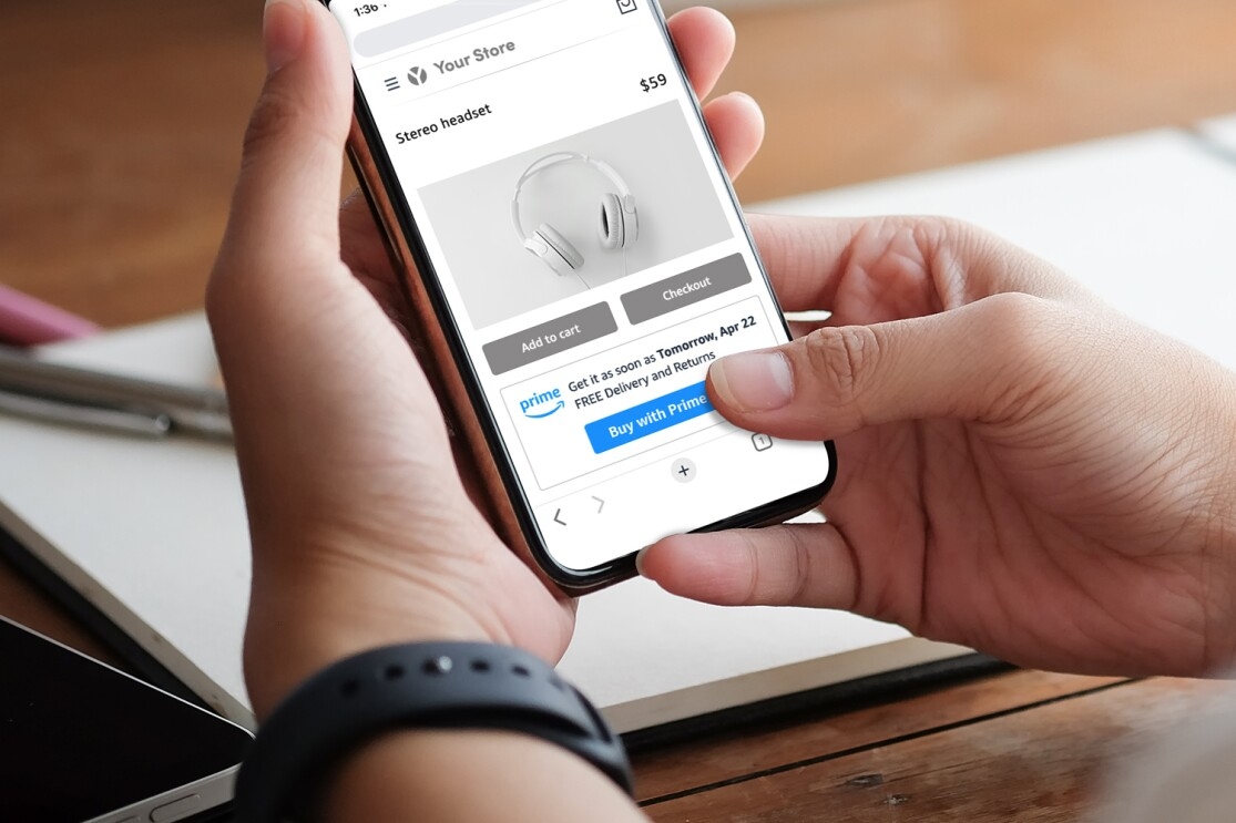 An image of two hands holding a phone. One the phone's screen, you see the process of someone buying headphones using Amazon's new "Buy it with Prime" feature on a merchant's website.