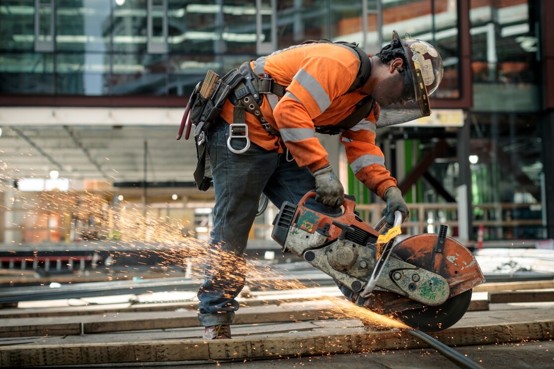 A man in a construction helmet and orange safety jacket is photographed at a construction site on Amazon's campus in Seattle, WA. He is using a circular saw to cut through metal. 