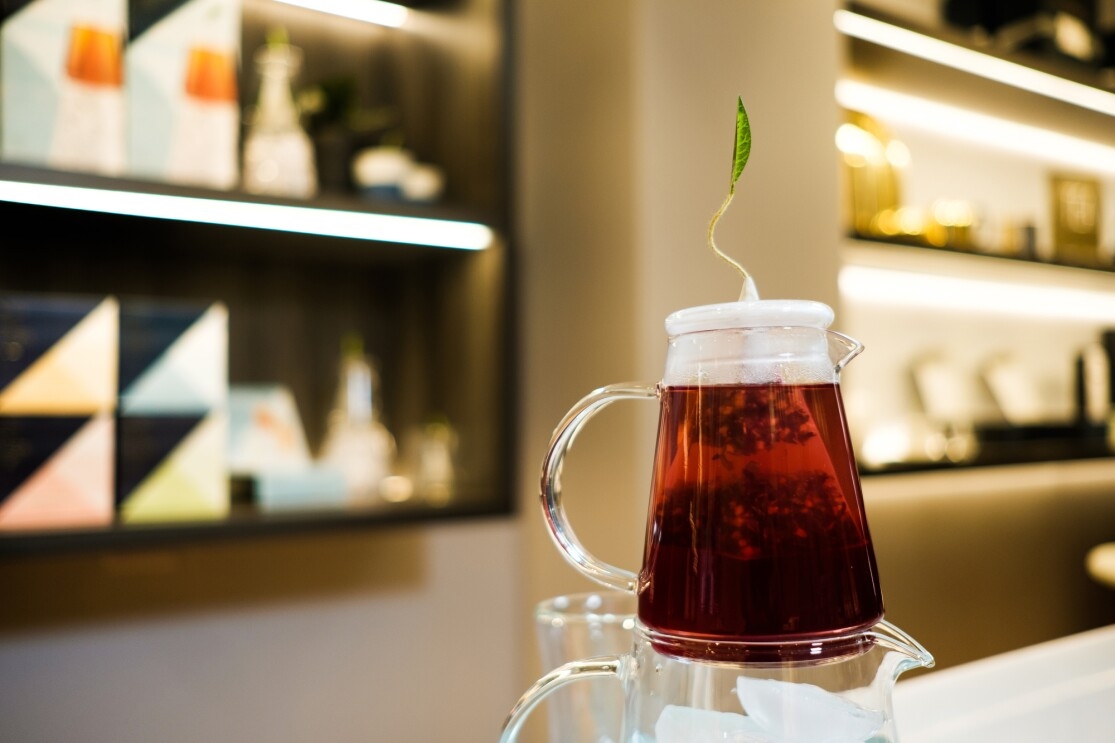 Tea steeps in a small glass pitcher adorned with a green leaf on top. The pitcher sits atop a larger pitcher filled with ice.