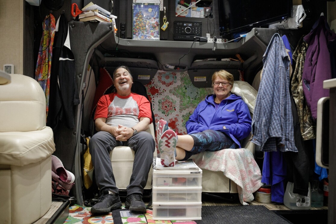 A man and woman sit in matching chairs inside their motorhome. The man is laughing, and the woman is smiling.